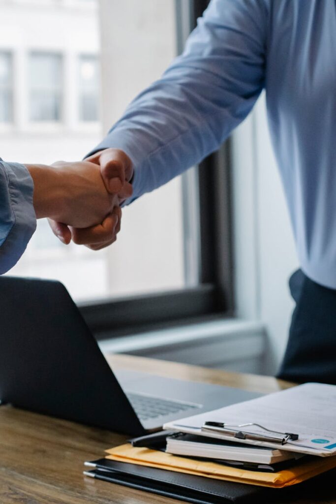Crop unrecognizable coworkers in formal wear standing at table with laptop and documents while greeting each other before meeting